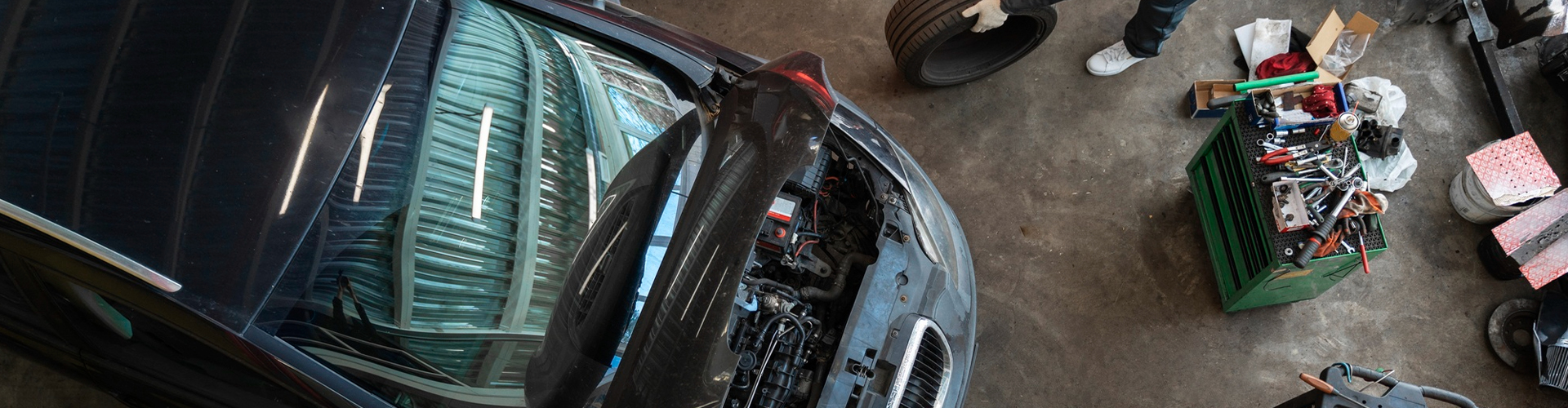 Top-down view of a car with its hood open in a workshop, engine exposed and a cluttered tool chest nearby