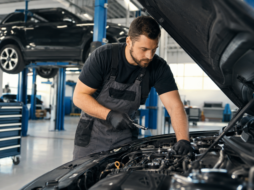 Mechanic in dark overalls inspecting the engine under an open hood in a bright auto repair shop with a car on a lift nearby.